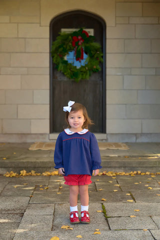 Toddler girl wearing Bon Air Bloomer Set in navy top with white collar, red bloomers, and embroidered bows standing outside a door.