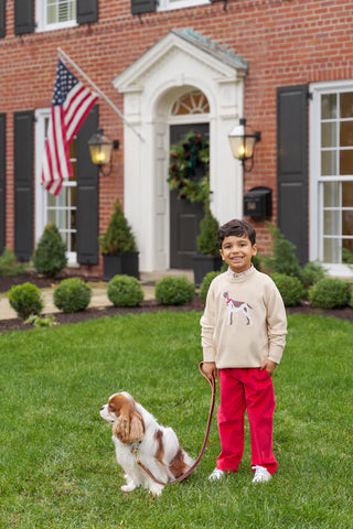 Boy wearing Isaac's Intarsia Sweater in osprey oatmeal with dog design, styled with red pants, standing outside a brick home with flag.