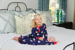 Little girl and baby wearing TBBC Sutton’s Sweet Dreams Pajamas in Jolly Old St. Nick print with Richmond Red trim on a white bed.