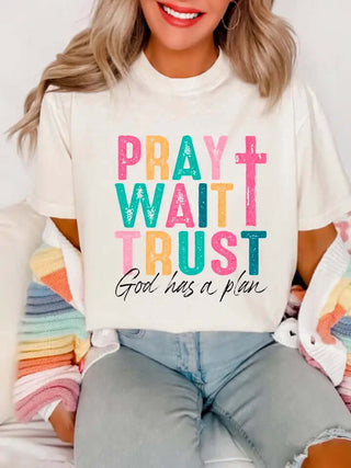 Pray Wait Trust Inspirational Tee—cream shirt with colorful block letters, cross graphic, and “God has a plan” faith message for women.