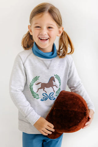Girl smiling in gray Cassidy Crewneck with horse and laurel design, holding brown equestrian cap and wearing blue turtleneck underneath