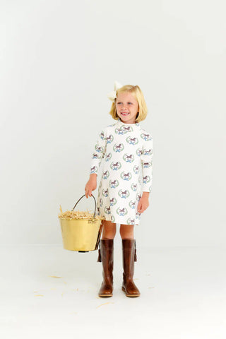 Smiling girl in Highland Horses turtleneck dress and tall brown boots, holding brass hay bucket against plain white background