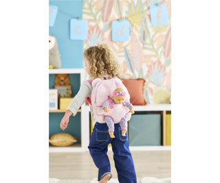 Toddler wearing pink plush Corolle backpack with 12-inch doll secured in front carrier straps while walking in playroom.