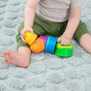 Baby playing on a soft rug with bright Dimpl Stack cups, holding multiple colorful pieces for stacking and sensory exploration.