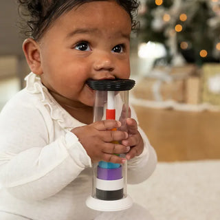 Baby holding a Fat Brain Flipty toy with colorful stacked rings inside clear tube while sitting on the floor and chewing on end piece