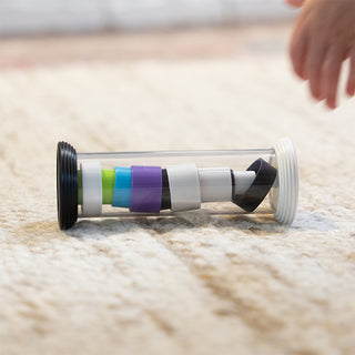 Flipty toy from Fat Brain lying on its side on the floor with rainbow colored and black and white rings stacked inside clear tube