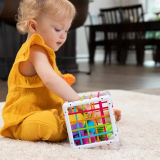 Toddler in yellow dress playing with Fat Brain InnyBin, pushing chunky textured pieces through elastic bands with curiosity