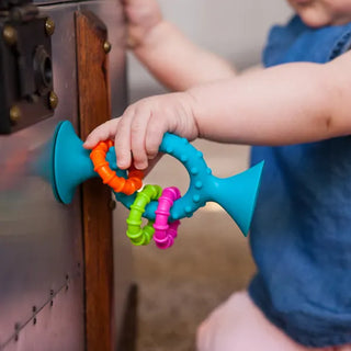 Close-up of blue pipSquigz Loops toy suctioned to a table, with baby’s hand exploring textured silicone handle and colorful rattling loops.