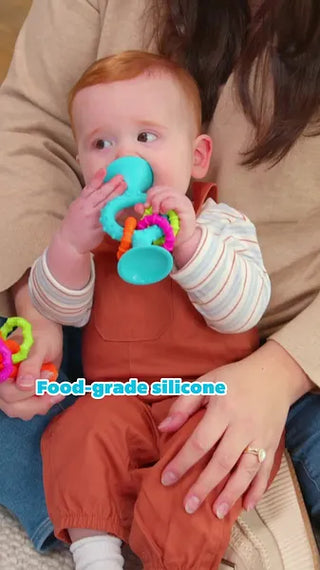Baby chewing on bright blue pipSquigz Loops teether toy with colorful rattle rings, made from food-grade silicone for safe sensory play.