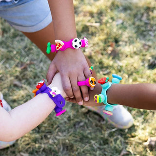 Three children show off their Plip Charms bracelets in the grass, featuring fun designs like pizza, animals, and strawberries.