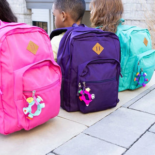 Plip Charms clipped to colorful backpacks—pink, purple, and aqua—adding fun, fidgety flair to school bags on the playground.