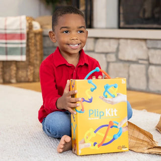 Boy in red shirt smiles while holding Plip Kit box with bright orange packaging and bendable sensory toy pieces shown on the front.