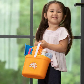 Happy girl proudly holds orange Pretendables Cleaning Kit bucket filled with toy cleaning tools near a window with natural light.