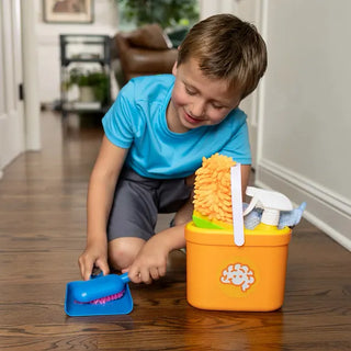Smiling boy uses brush and dustpan from Pretendables Cleaning Kit while caddy holds mop, towel, spray bottle, and other tools beside him.