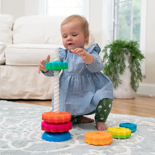 Baby girl in blue dress stacking vibrant Fat Brain SpinAgain discs on white corkscrew pole while sitting on living room rug.