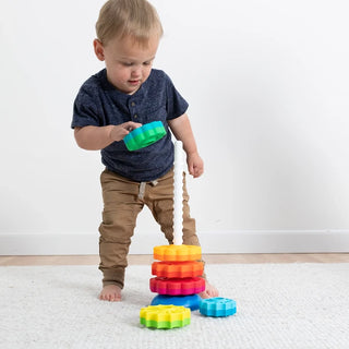 Toddler holding green disc, playing with colorful SpinAgain stacking toy on carpeted floor with white wall background.