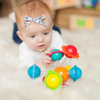 Baby on tummy time reaching for Fat Brain Wimzle sensory toy with colorful textured balls and silicone rings for teething and play.