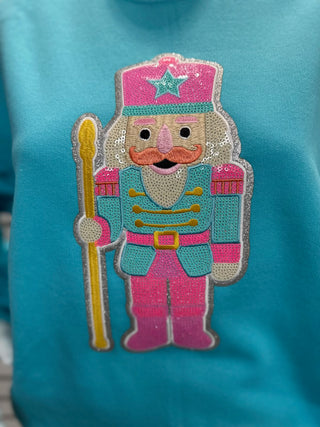 Close-up of women’s aqua sweatshirt highlighting the sparkling sequin nutcracker patch with intricate stitching and bold colors.