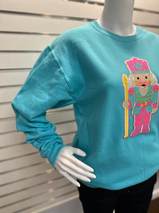 Women’s aqua sweatshirt on mannequin featuring colorful sequin nutcracker patch with festive detailing and soft crewneck fit.