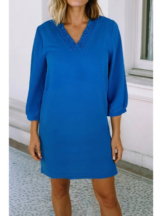 Woman wearing blue ruffle V-neck dress with three-quarter sleeves and a relaxed, flowy fit, standing near a white building.