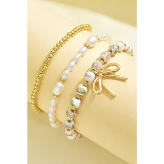 Close up of three stacked beaded bracelets featuring gold beads, pearl beads, floral beads, and a gold bow charm