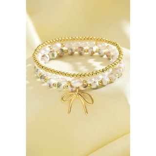 Triple layer beaded bracelet set with gold beads, pearls, floral beads, and gold bow charm displayed on soft yellow fabric