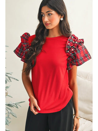 Woman wearing a red holiday tee with plaid ruffle sleeves and pearls, standing by a Christmas tree in a festive indoor setting.