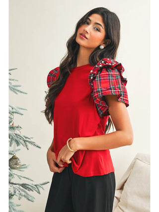 Side pose of woman wearing a red tee with plaid ruffle sleeves decorated with pearls, styled by a tree for the holiday season.