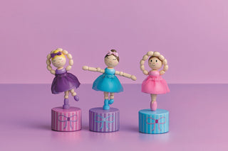 Three wooden collapsing toy ballerinas in colorful pink, purple, and blue dresses on cylindrical stands against a pink background