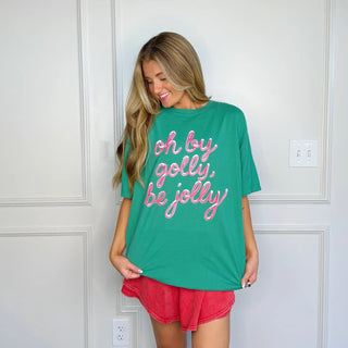 Woman wearing green Oh By Golly Be Jolly Christmas tee paired with red shorts, standing indoors and smiling.