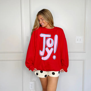 Woman wearing bright red JOY graphic Christmas sweatshirt with polka dot shorts, smiling and posing in front of a wall.