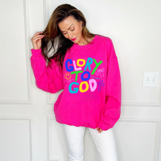 Woman wearing bright pink Glory to God sweatshirt with colorful lettering standing against white wall in casual faith-inspired outfit.