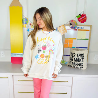 Woman wearing cream Happy Teacher Club sweatshirt with colorful playful school-themed graphics in a bright classroom setting.