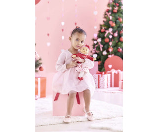 Child holding Corolle Bebe Calin Margaux Heart Petals doll in pink holiday setting with decorated tree and gifts.